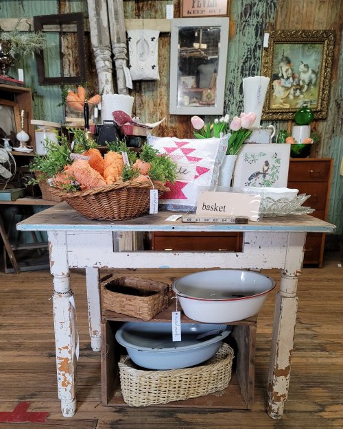 A rustic table displays a basket of carrots, flowers, and decorative items in a charming vintage shop setting.