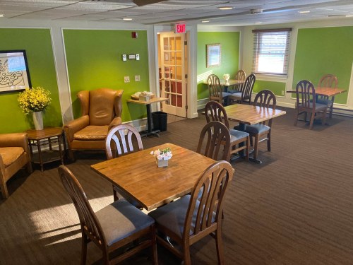 Cozy dining area with wooden tables, chairs, and green walls, featuring a flower arrangement and natural light.