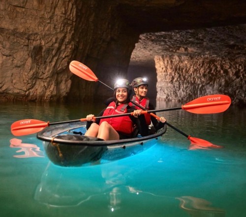 Two people in a kayak with orange paddles, exploring a cave with glowing blue water and headlamps on.