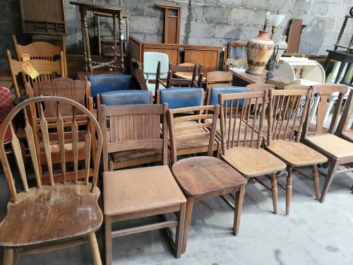 A variety of wooden chairs in different styles and conditions, arranged in a warehouse setting.