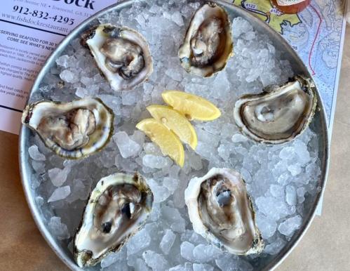 A metal platter filled with ice and six fresh oysters, garnished with lemon wedges.