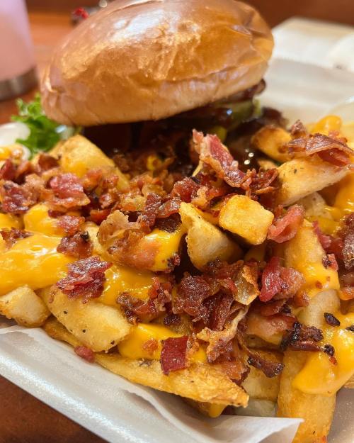 A close-up of loaded fries topped with cheese and crispy bacon, alongside a burger in the background.