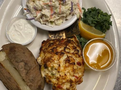 A plate featuring crab cake, baked potato, coleslaw, lemon wedge, and a side of honey.