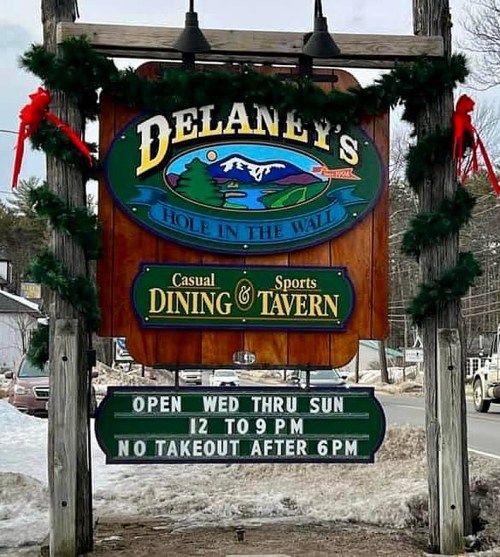 Sign for Delaney's Hole in the Wall, a casual dining and sports tavern, with holiday decorations and hours listed.