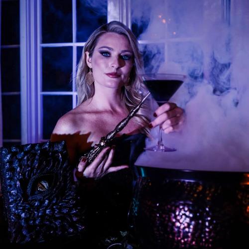 A woman in a dark outfit holds a cocktail and a wand, surrounded by smoke and eerie lighting.
