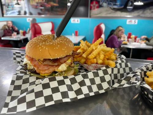 A close-up of a burger with bacon and fries on a checkered tray, with a diner setting in the background.