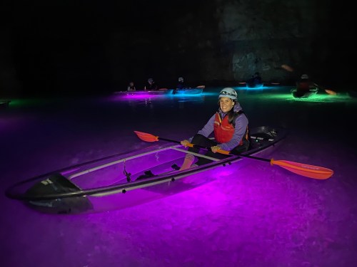 A person in a kayak with orange paddles, surrounded by glowing purple water in a dark cave.