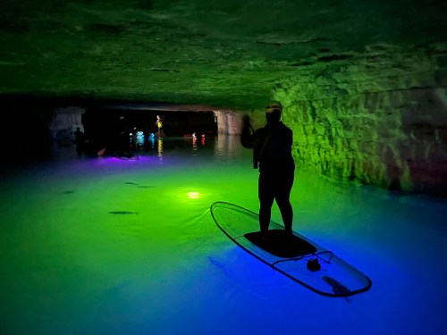 A person stands on a paddleboard in a colorful, illuminated cave with glowing water and rock formations.