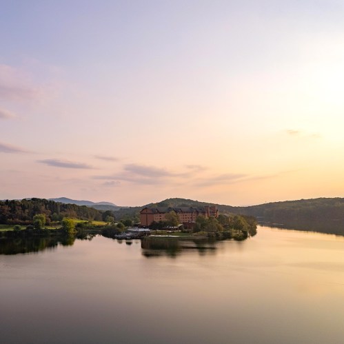 A serene landscape featuring a calm lake, lush greenery, and a distant building under a pastel sunset sky.