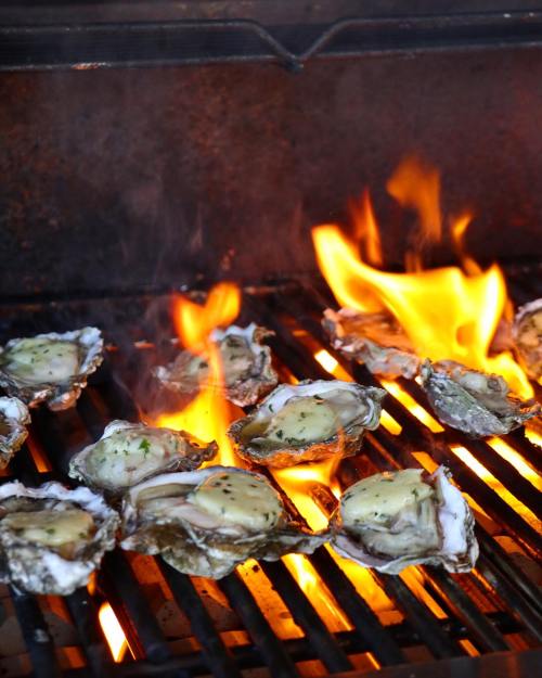 Grilled oysters on a barbecue grill with flames and smoke rising, showcasing a delicious cooking process.