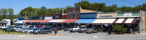 A charming small-town street lined with shops, colorful awnings, and festive decorations, with parked cars in front.