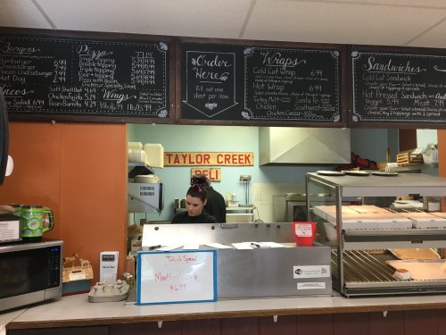 A deli counter with a menu board, a worker preparing food, and a sign reading "Taylor Creek Deli."