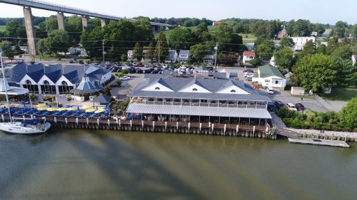 Aerial view of a waterfront restaurant with outdoor seating, surrounded by greenery and parked cars.