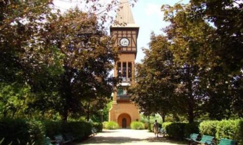 A tall clock tower surrounded by lush trees and benches in a park setting on a sunny day.