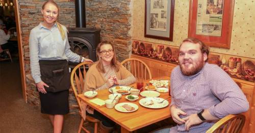 A waitress stands beside two diners at a table with plates of food in a cozy restaurant setting.