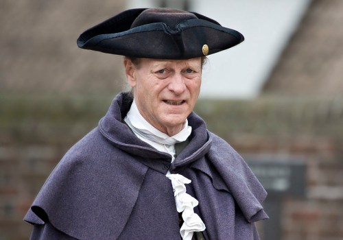 A person in historical attire, wearing a black hat and a purple cloak, stands outdoors with a neutral expression.