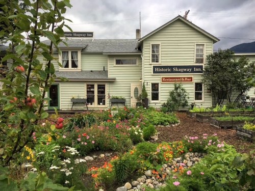Historic Skagway Inn with a vibrant flower garden in front, set against a cloudy sky.