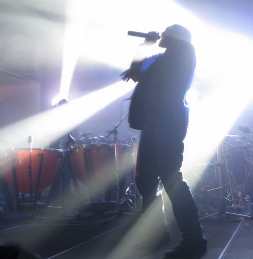 A silhouetted performer sings into a microphone, surrounded by dramatic lighting and percussion instruments.
