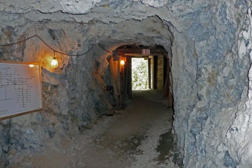 A dimly lit mine tunnel with rocky walls, featuring exit signs and a whiteboard on the left.