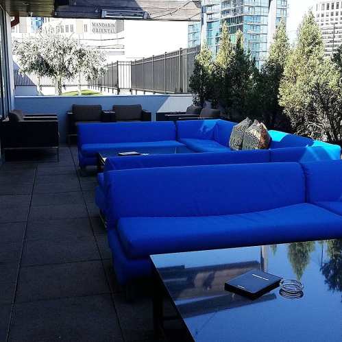 A modern outdoor lounge area with blue couches and a glass table, surrounded by greenery and city buildings.