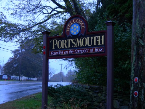 Welcome sign for Portsmouth, founded in 1638, with a rainy backdrop and trees lining the road.