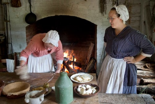 Two women in historical attire work in a rustic kitchen, preparing food by a fireplace with various dishes on the table.