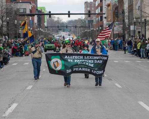 A parade scene with participants holding a banner for the St. Patrick's Day Parade in Lexington, surrounded by a cheering crowd.
