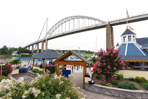 A scenic waterfront view featuring a bridge, restaurant, and blooming flowers along the riverbank.