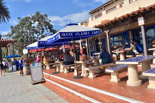 Outdoor dining area of Greek Islands Café with blue umbrellas, people enjoying meals, and a sunny atmosphere.