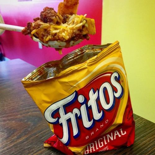 A partially open bag of Fritos with a spoonful of chili and cheese lifted above it, resting on a wooden table.