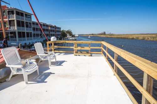 A sunny deck with two white chairs overlooking a calm waterway and marshland, with buildings in the background.