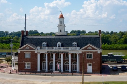 Historic brick building with a clock tower, overlooking a river, under a blue sky with scattered clouds.