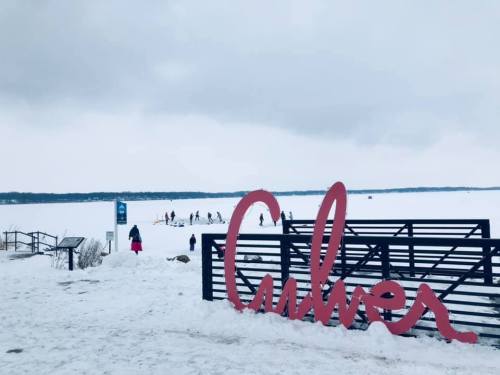 A snowy landscape with a large "Culver" sign in the foreground and people ice fishing on a frozen lake in the background.