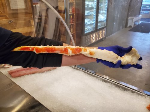 A person wearing blue gloves holds a large crab leg over a bed of ice in a seafood market.