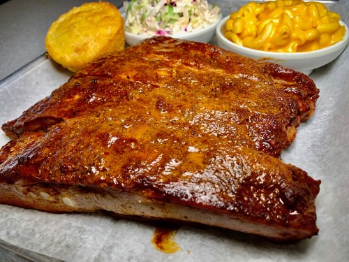 A plate featuring a large portion of glazed ribs, coleslaw, macaroni and cheese, and cornbread muffin.
