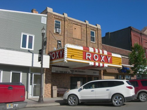 Historic Roxy Theater marquee in a small town, with a red and yellow sign, surrounded by buildings and parked cars.