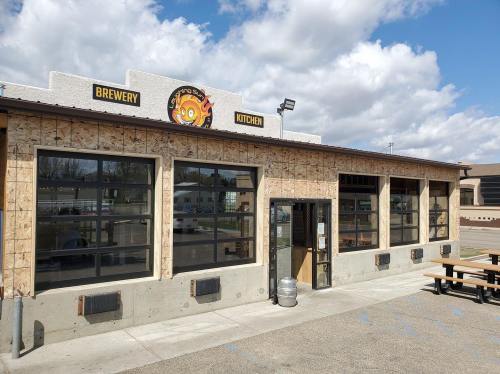 Exterior of a brewery and kitchen with large windows, a sign, and picnic tables outside under a partly cloudy sky.