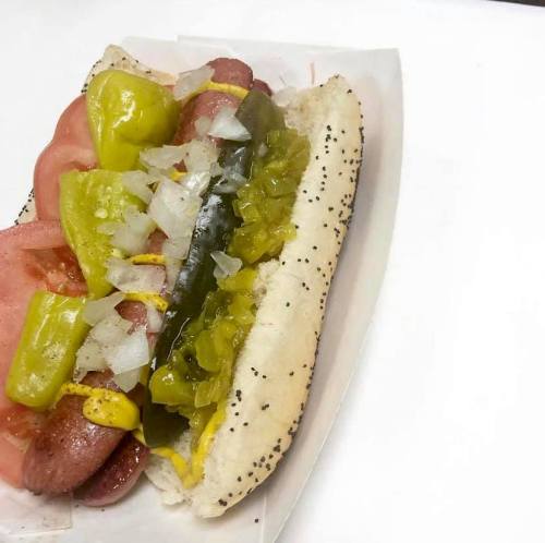 A Chicago-style hot dog in a poppy seed bun, topped with tomatoes, pickles, onions, and yellow mustard.