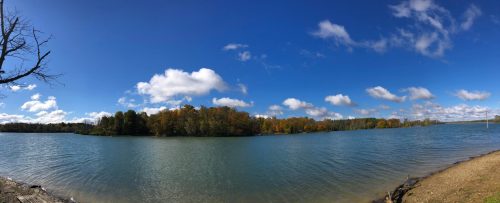 A panoramic view of a serene lake surrounded by trees with autumn foliage under a bright blue sky and fluffy clouds.
