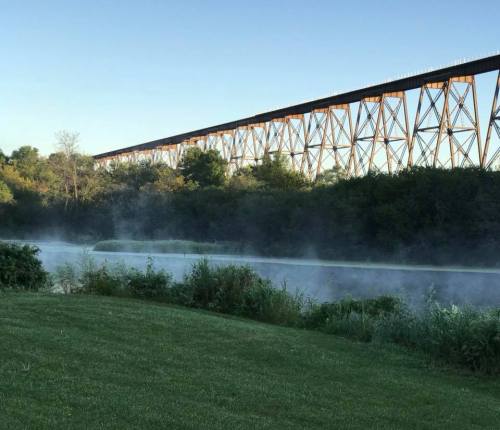 A misty river scene with lush greenery and a tall, rust-colored bridge in the background under a clear blue sky.