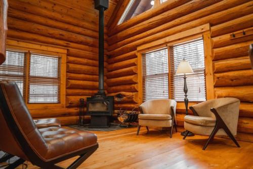 Cozy log cabin interior with wooden walls, two armchairs, a lamp, and a wood stove. Natural light from windows.