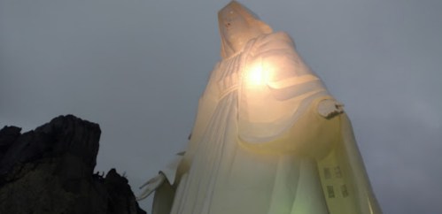 A towering statue of a figure illuminated against a cloudy sky, surrounded by rocky terrain.