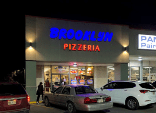 A brightly lit pizzeria sign reading "Brooklyn Pizzeria" at night, with cars parked outside.