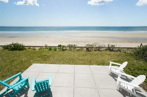A serene beach view with a clear sky, featuring a patio with colorful chairs overlooking the sandy shore.