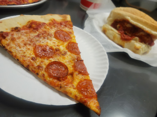 A slice of pepperoni pizza on a paper plate next to a sandwich filled with meat and sauce, on a table.