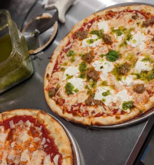 Two pizzas on metal trays: one topped with sausage and pesto, the other with tomato sauce and cheese.