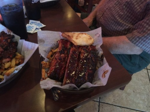 A plate of BBQ ribs with fries and a piece of bread, sitting on a table next to a person.
