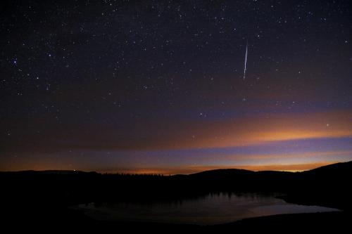 A starry night sky with a shooting star above a calm lake and silhouetted hills in the foreground.