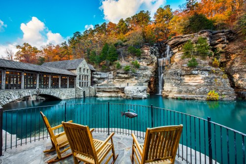 Scenic view of a tranquil lake with a waterfall, surrounded by autumn foliage and a stone building, featuring wooden chairs.
