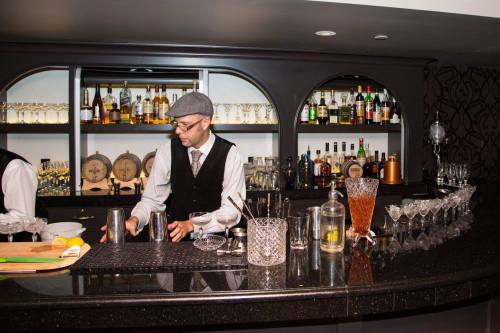A bartender in a vest and cap mixes drinks at a stylish bar with shelves of liquor and glassware in the background.